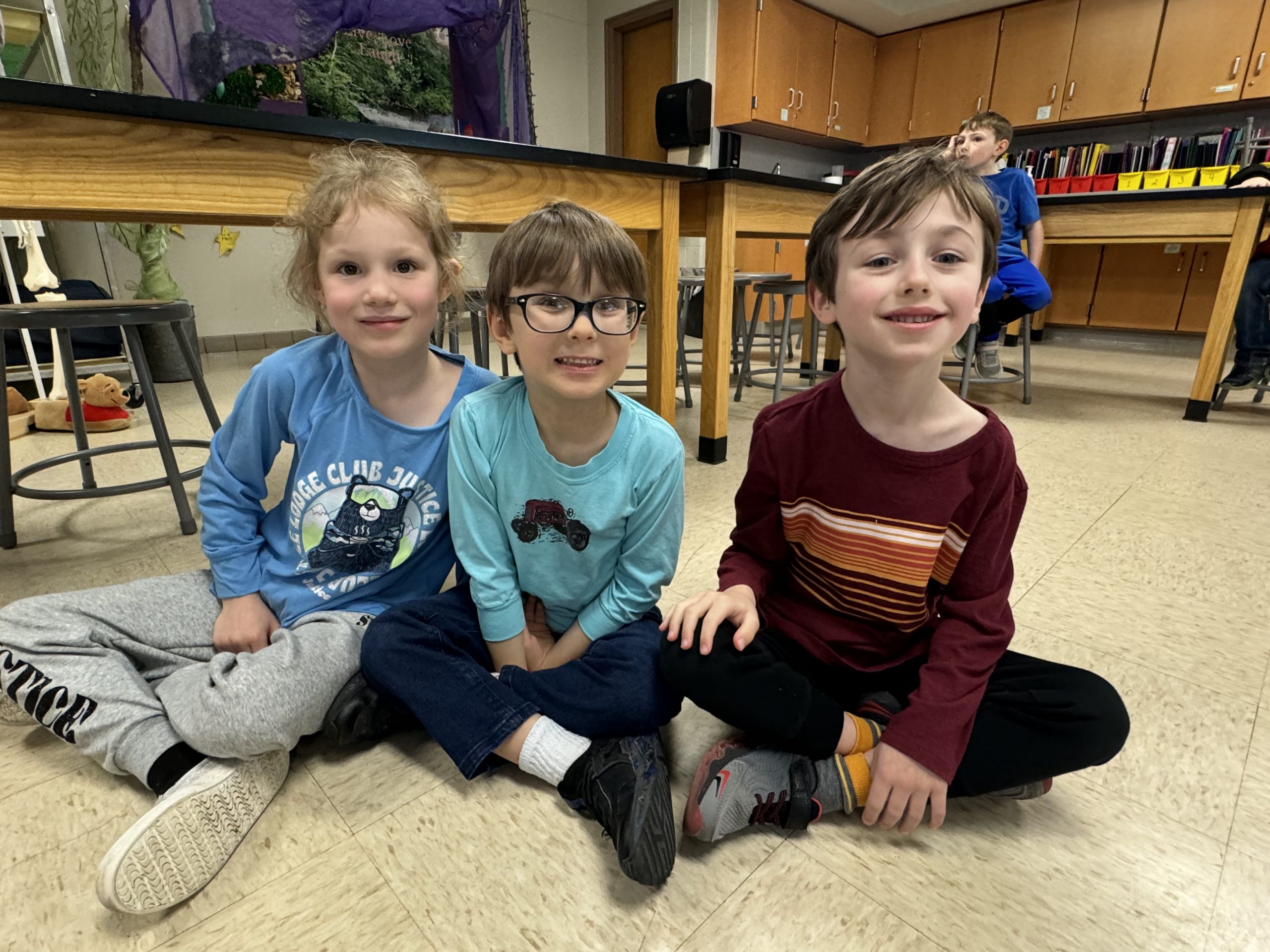 Three elementary school students sit on floor cross-legged with smiles